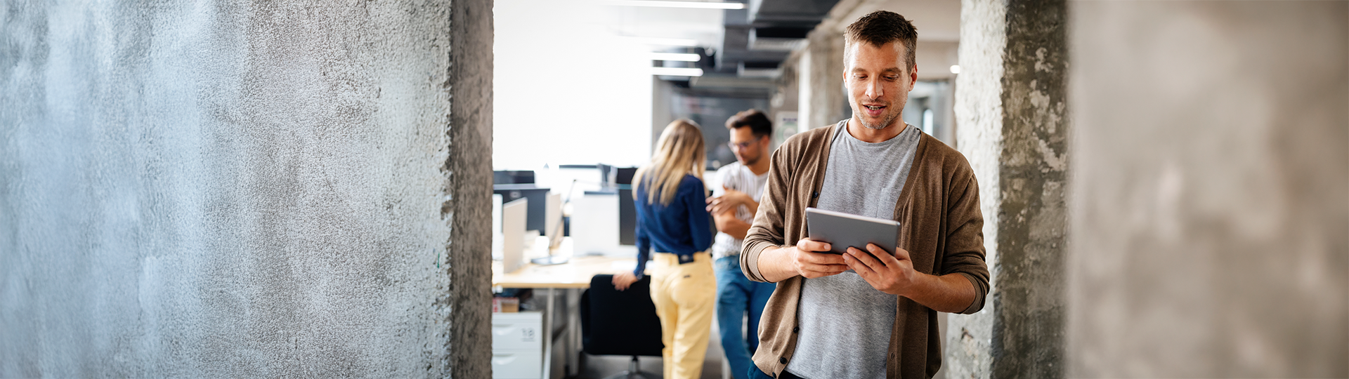 Um homem em um escritório moderno com estilo industrial utiliza um tablet enquanto caminha. Ao fundo, colegas de trabalho conversam em um ambiente colaborativo. A cena ilustra como o investimento em mdm permite que colaboradores utilizem dispositivos móveis com segurança e produtividade no dia a dia da empresa.