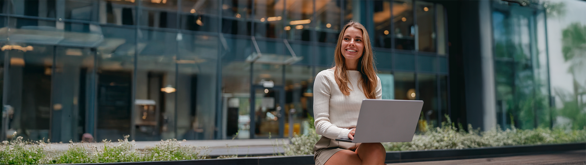 Uma mulher jovem e sorridente sentada em um ambiente externo moderno, utilizando um notebook no colo. Ao fundo, a fachada de um prédio comercial de vidro. A imagem ilustra a flexibilidade e segurança proporcionadas por uma eficiente gestão de MDM no ambiente corporativo.