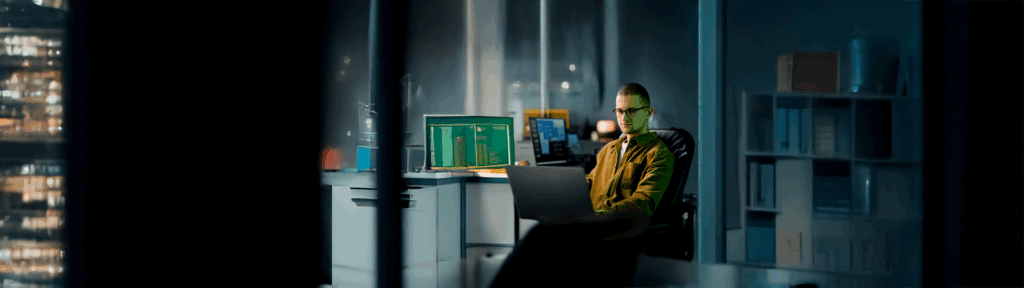 Um profissional de TI trabalhando em um escritório moderno à noite, analisando dados em um notebook e monitores. A imagem ilustra o processo de ITAD (Asset Disposition), focando na gestão segura e estratégica de ativos tecnológicos de uma empresa.