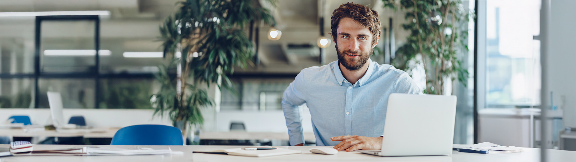 Homem jovem de camisa azul clara sorri em frente a um notebook aberto em um escritório moderno. Ao fundo, plantas decorativas e outras estações de trabalho representam o ambiente de uma empresa de tem.