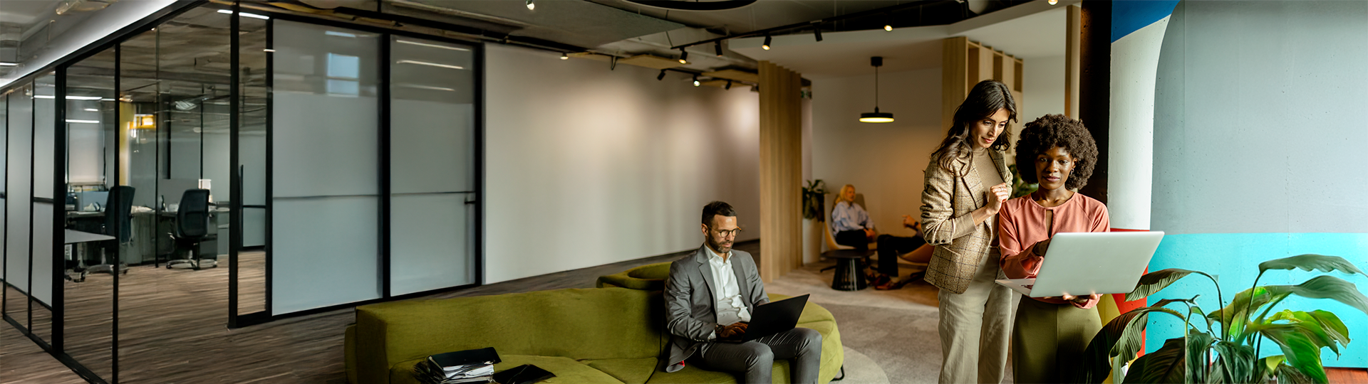 Uma foto panorâmica de um escritório moderno e colaborativo, ideal para ilustrar soluções de TEM, ITAM e MDM. No lado direito, duas mulheres analisam juntas a tela de um notebook. No centro, um homem de terno trabalha concentrado em seu laptop, sentado em um sofá verde oliva. Ao fundo, outras pessoas conversam em uma área de convivência, enquanto o lado esquerdo exibe salas de reunião com divisórias de vidro, destacando um ambiente corporativo tecnológico e organizado.